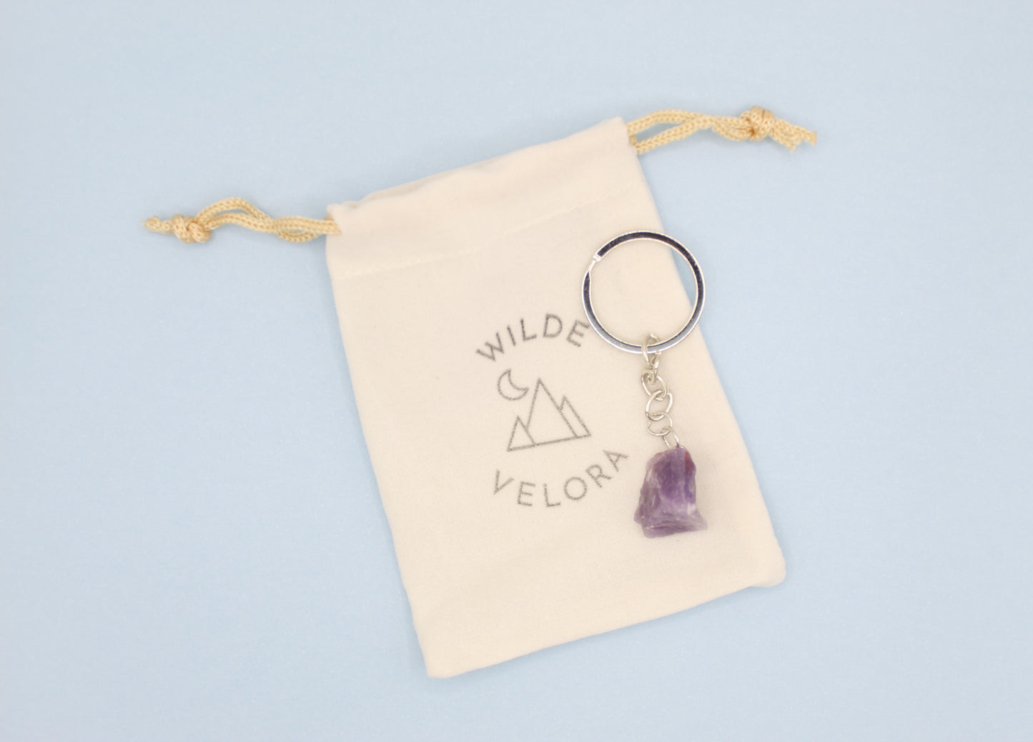 Keychain with purple crystal on a beige drawstring bag with 'Wilde Velora' branding on a light blue background.