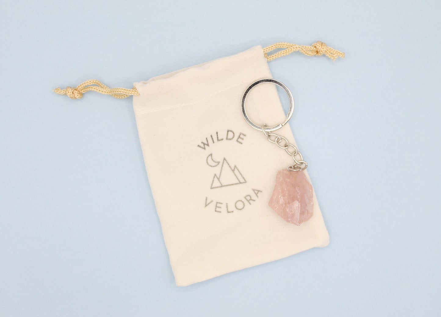 Keychain with pink stone and gold chain on a beige drawstring bag with 'Wilde Velora' branding, on a light blue background.