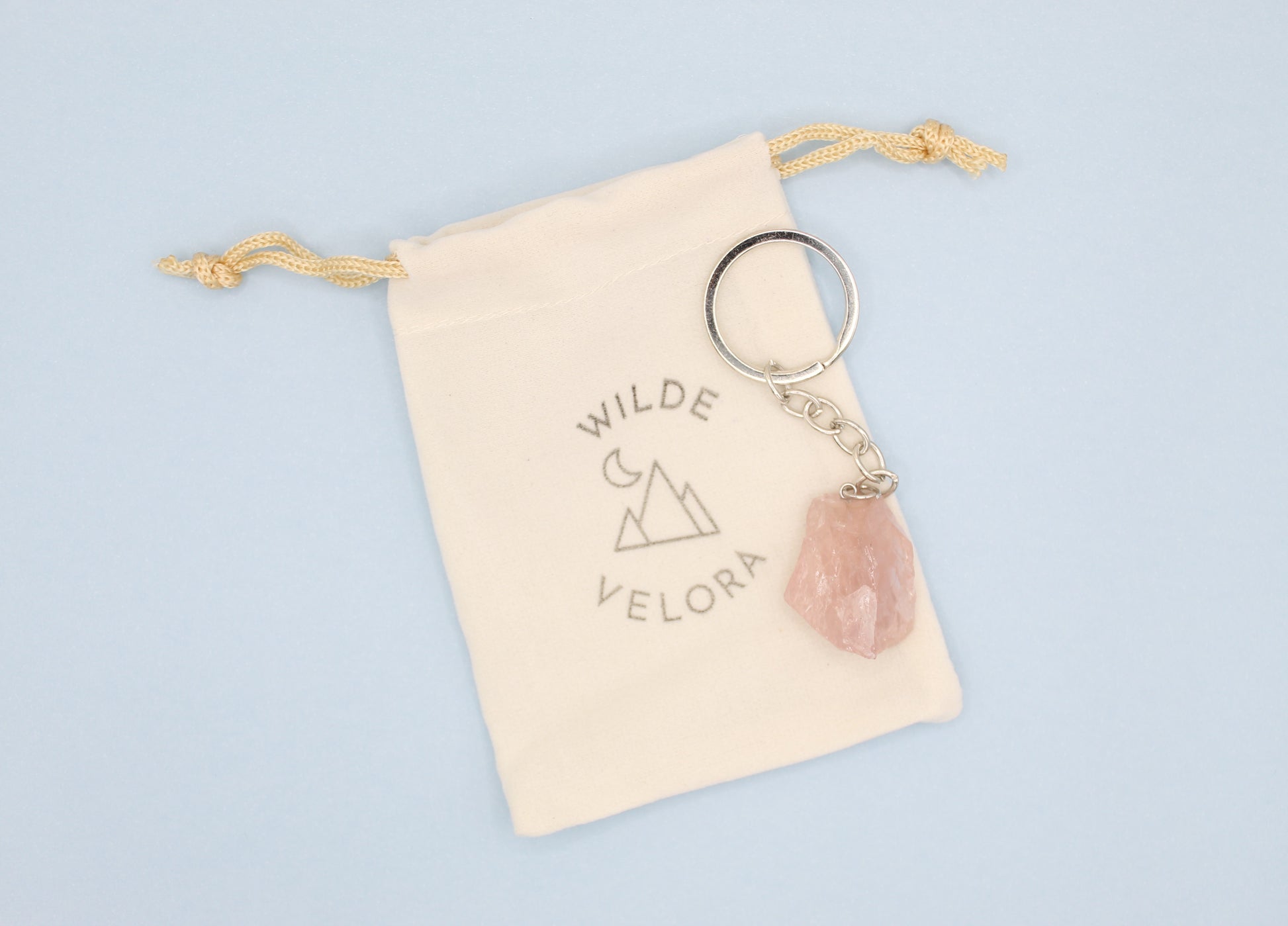 Keychain with pink stone and gold chain on a beige drawstring bag with 'Wilde Velora' branding, on a light blue background.